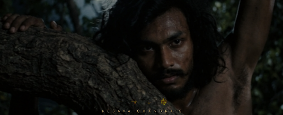 Actor Kesava Chandra as AGENETOS standing in a dark forest with an intense expression, in a still from the upcoming epic film LUCIFERUM, part of the PROJECTUM 000° cinematic universe.